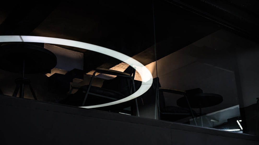 White LED pendant light, suspended above the dining table in an office building with black chairs and dark walls, in a minimalist style, closeup of circular frame lens, dim lighting, soft tones, and a sense of space. The reflection on the glass wall creates depth and perspective. in the style of minimalism.