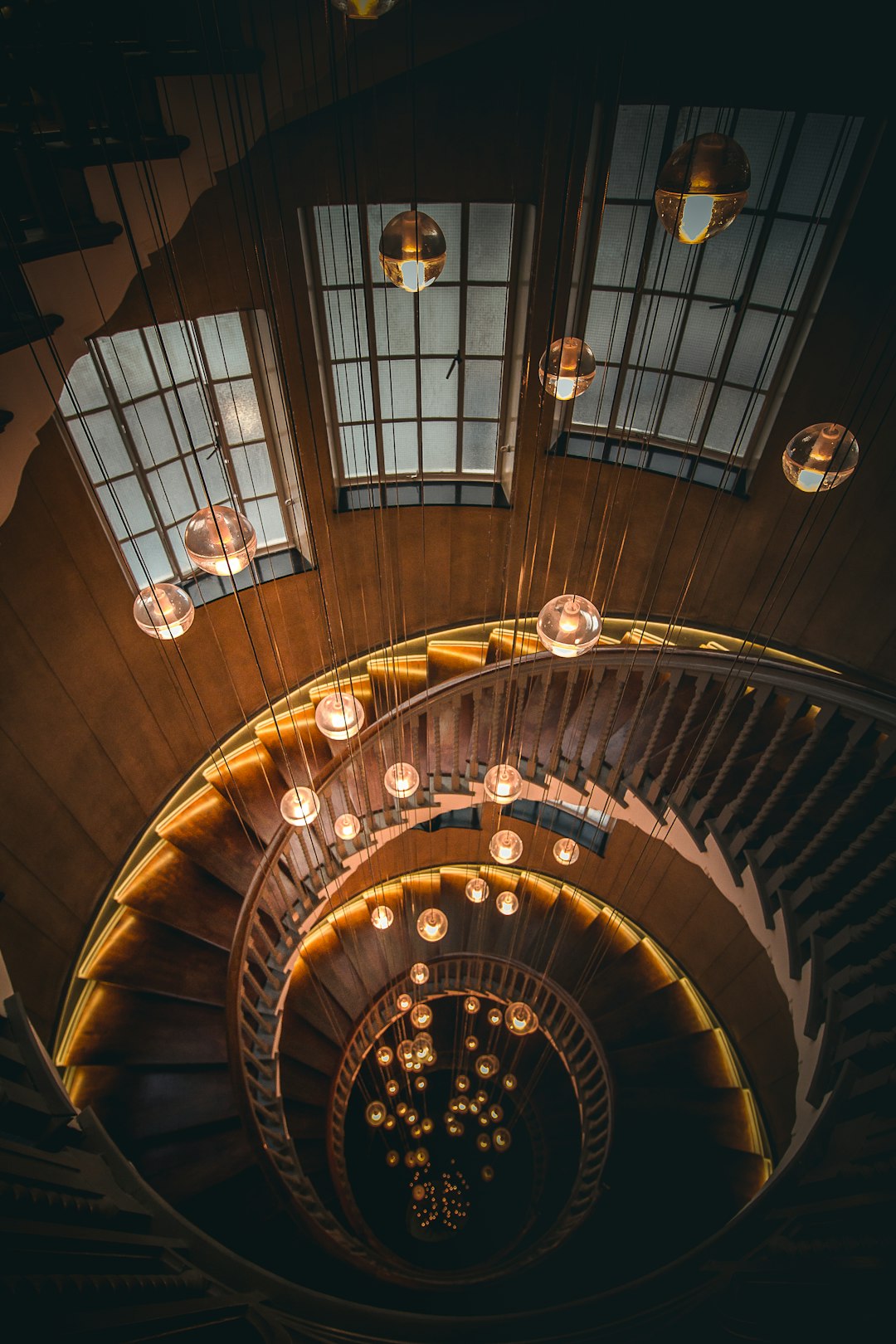 In the center of an old wooden staircase, several glass chandeliers hang from above, casting light on the spiral stairs below. The walls and ceiling have dark tones with light wood accents, creating a warm atmosphere. In contrast to the lights hanging down, there is no one in sight, giving it a lonely feel. This photo was taken using a Canon eos r5, with a focal length of f/20, aperture wide open, and shutter speed long exposure time, capturing every detail of each lamp’s design in the style of long exposure photography.