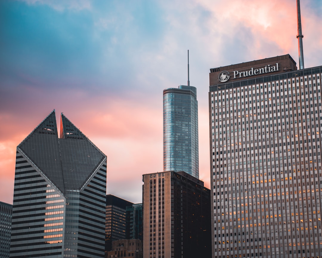 A photograph of the “PrudPortable” logo on top of a building in Chicago, with iconic skyscrapers like John Hancock and super tall buildings around it. The sky is pastel pink and blue at dusk, creating a beautiful sunset backdrop. In soft focus behind them can be seen famous landmarks such as Millennium Park’s bean sculpture or the Michigan Avenue Bridge. Captured in the style of Nikon Z6 II camera using a wide-angle lens, with natural lighting. –ar 64:51