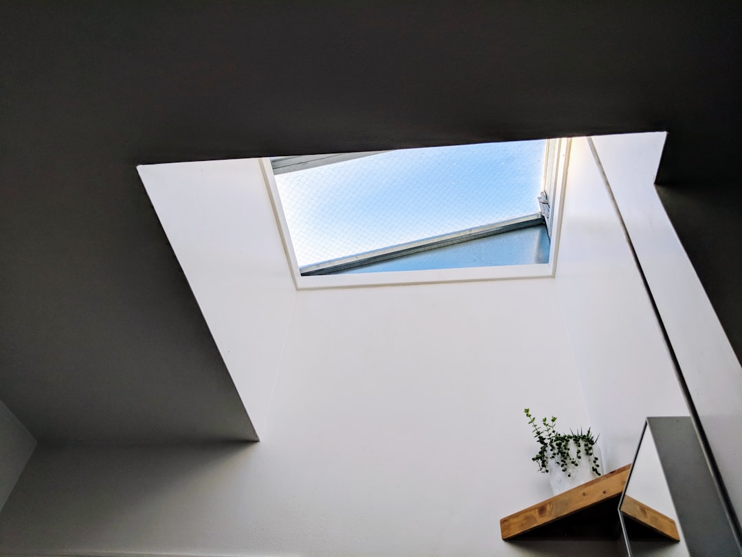 A skylight in the ceiling of an apartment, white walls, grey background, minimalist interior design photography, window on one side, small desk with plants underneath it, skimming light coming through the skylight, shot from below, angled view. –ar 4:3