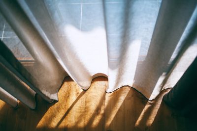 A closeup of white curtains on the window, with sunlight shining through them and casting long shadows on the wooden floors below. The scene is captured in a photo taken from above in the style of Hasselblad X2D 50C camera with Portra Pro 400 film. It has a soft focus and a natural color palette.