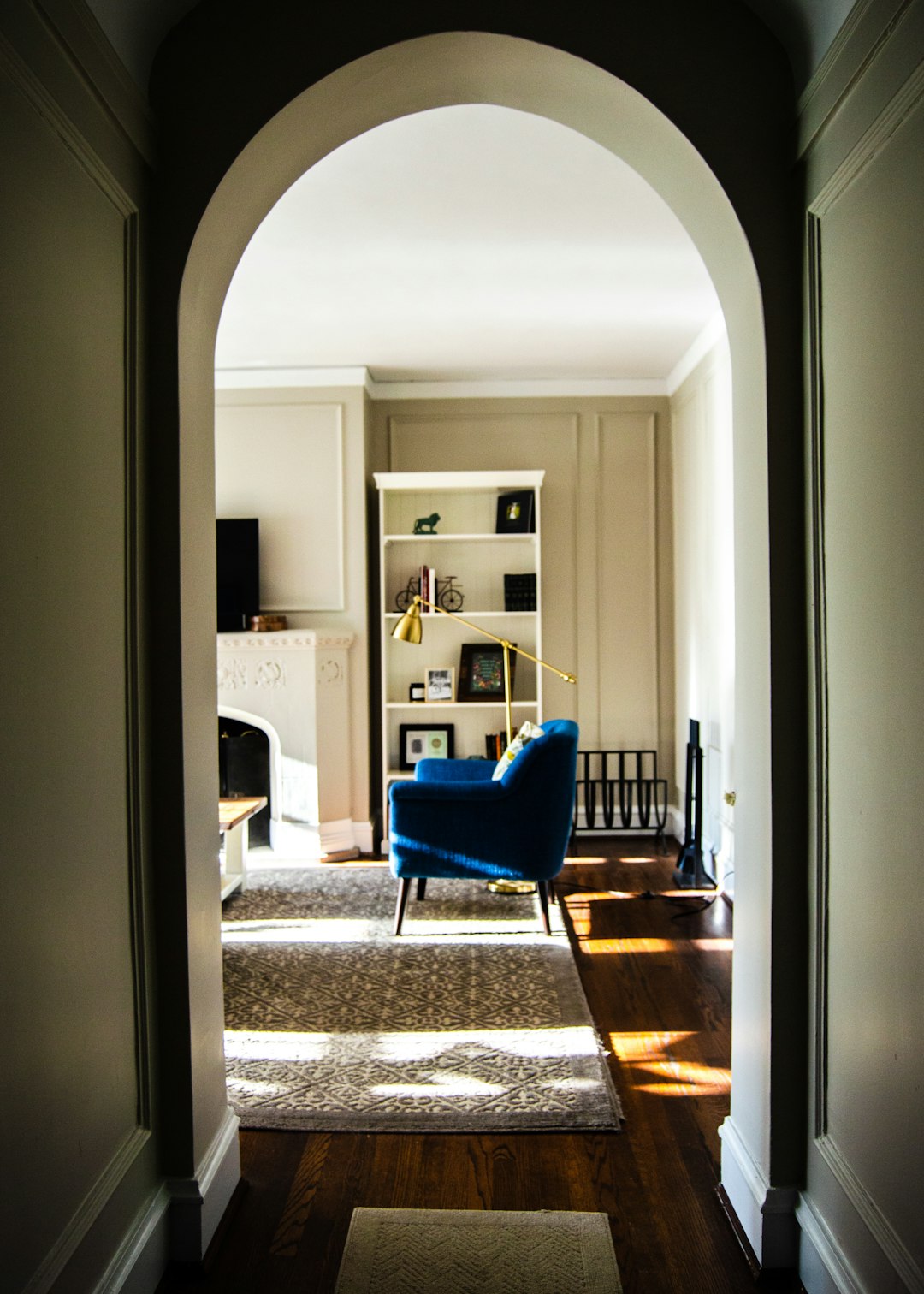 The view from the doorway of an arched wall leading to a living room with a blue chair, rug and white bookcase. Use a Canon EOS camera for high-resolution images with natural lighting. The perspective should be straight-on, capturing details like soft shadows and warm tones in each corner of the home.