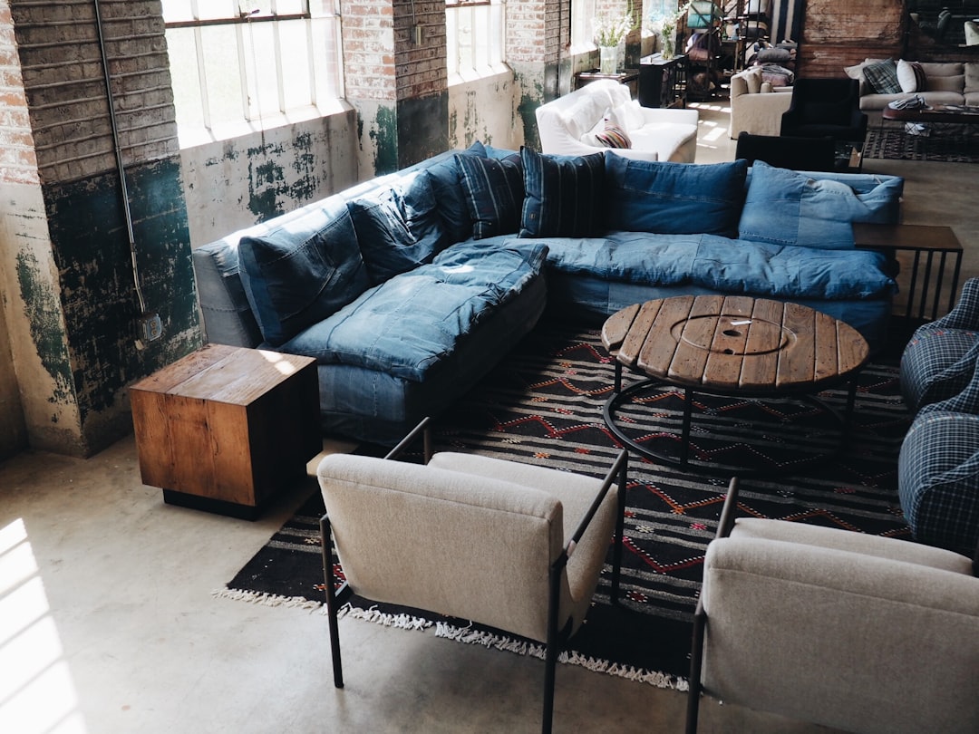 A large, comfortable blue denim sofa in an industrial-style living room with rustic brick walls and vintage furniture. The scene is captured from above in the style of a wide-angle Canon EFS f/2.8mm lens, creating a warm atmosphere. This photo was taken using Fujifilm Provia film, adding warmth to its surroundings. It features high resolution and detailed textures, showcasing the unique texture of jeans with minimal editing of the original text.