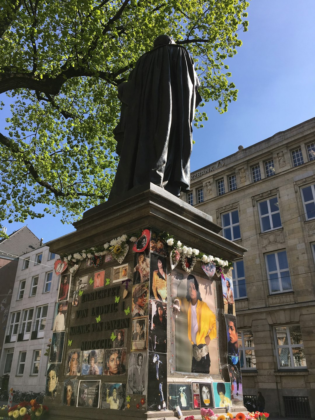 A photo of the monument to Michael Jackson in Hamburg, Germany. The statue is covered with photographs and flowers from fans who come every day. It stands on top of an old building in front of a blue sky. A large tree sits behind it. In the background you can see other buildings. Bright sunlight shines through green leaves onto parts of the sculpture. At some places around the base, people have placed mementos for him. Some images show that he was surrounded by adoring fans in the style of adoring fans. –ar 3:4