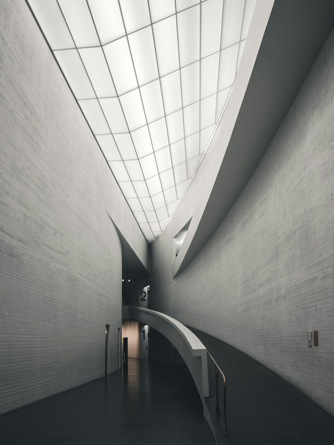 The interior of the museum has a ceiling and walls made of white concrete. An overhead skylight illuminates one side of the wall with a light gray and black color palette. A long curved staircase leads to the second floor in front, with architectural photography in the style of hyper realistic.