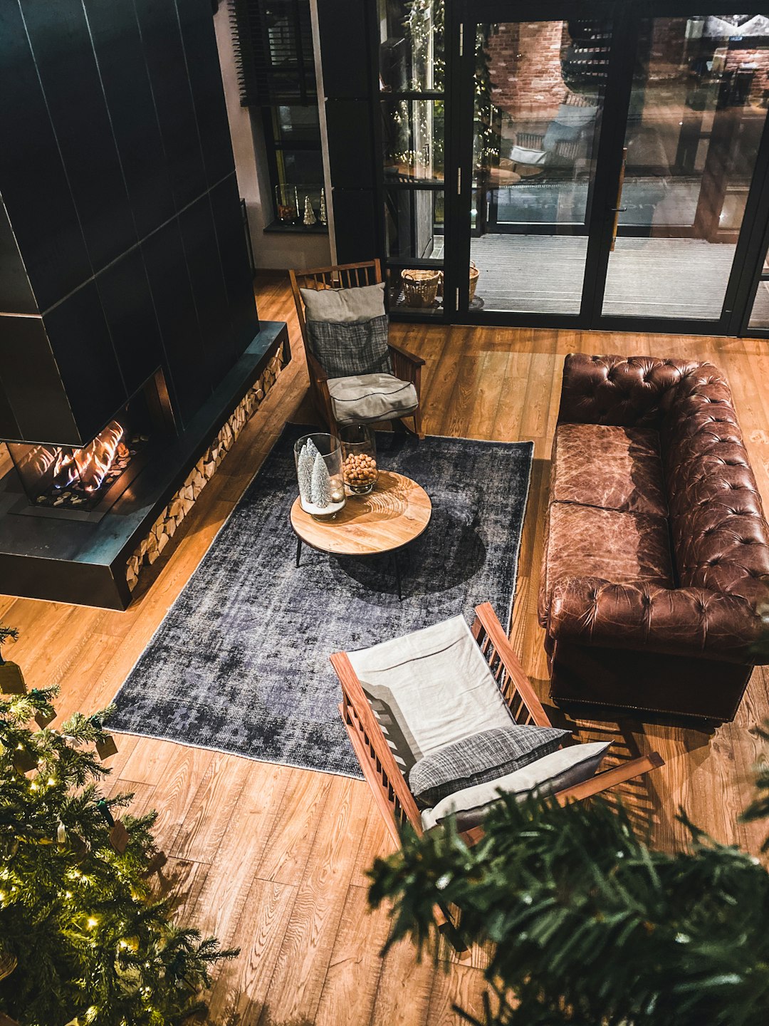 A cozy living room with leather sofas, wooden floors and black fireplace in an industrial style home, seen from above. The scene includes a grey rug on the floor, wooden flooring, large windows, a coffee table adorned with stylish decor, potted plants, and a plush brown couch, creating a warm atmosphere.