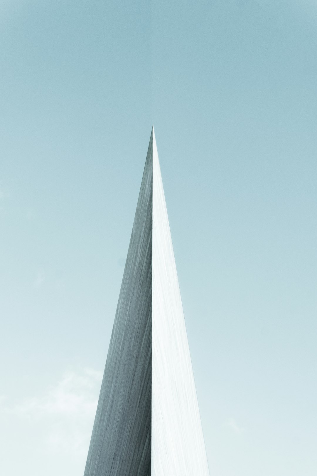 A minimalist photo of an architectural structure with sharp edges, standing against the clear sky. The shape is reminiscent of a triangle or pyramid, with its long side facing upwards and positioned towards the top left corner of the frame. It is white in color, creating contrast with the light blue background. This composition creates depth by emphasizing perspective through a subtle angle on one end while capturing a sense of grandeur from a low angle view, in the style of minimalism. –ar 85:128