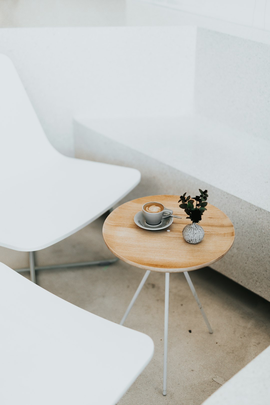 A small round wooden table with white legs and an open cup of coffee on it, placed in the corner between two simple modern chairs and concrete walls, top view, minimalist style, natural light, neutral tones, clean lines, interior design photography, calm mood. The scene has a coffee shop vibe and boho feel, in the style of neutral colors. –ar 85:128