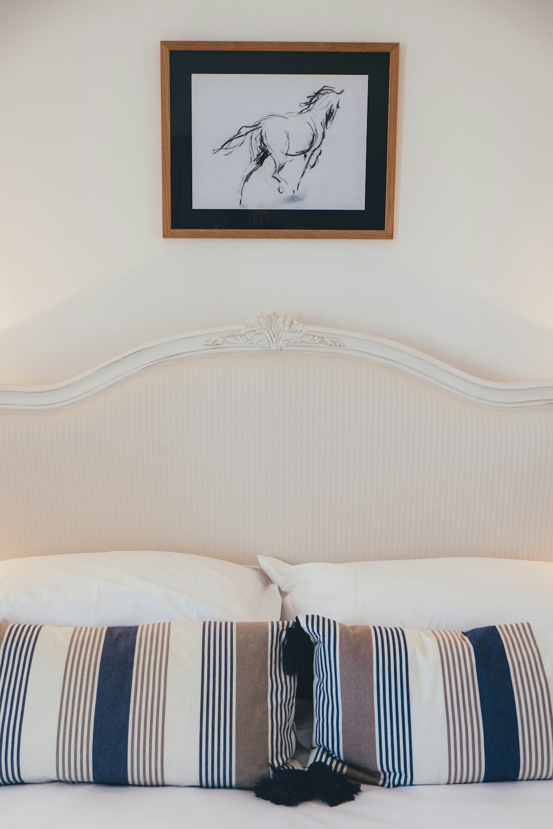 Close up of the headboard on a bed with white and navy blue striped pillows, in front there is one framed drawing with a horse sketch drawn in the style of hand hanging above the pillows, white wall background, closeup shot, professional photography with studio lighting. –ar 85:128