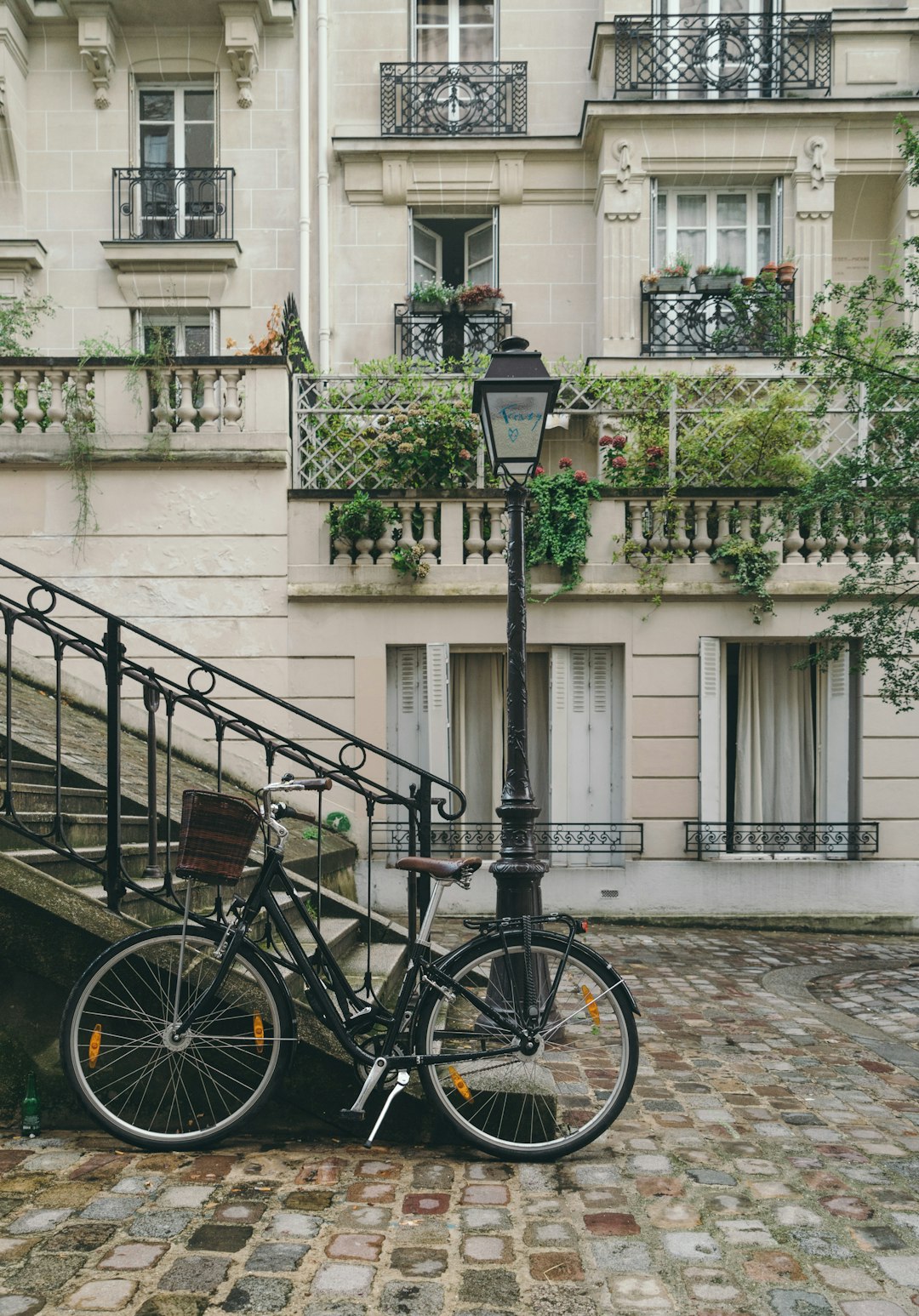 photo of a bike leaning against an old street light in front of apartment building with stairs leading up to the door, neutral tones, greenery hanging from windows and balconies, cobblestone ground, romantic Paris vibe –ar 89:128