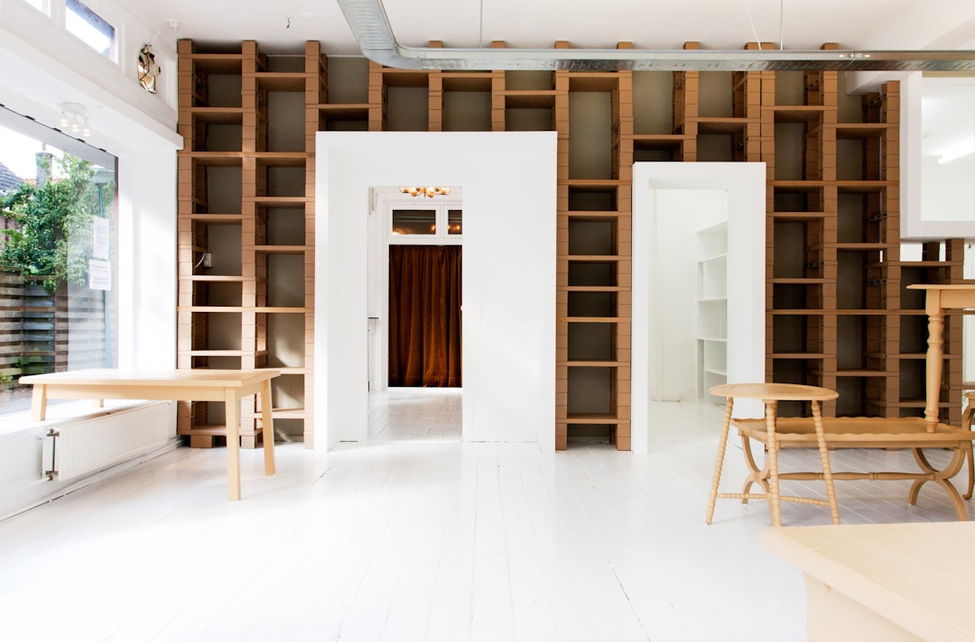 a photo of an open space with white painted floor, wooden walls and shelves, one door is closed on the left side and two doors in different sizes stand next to each other on right wall, there’s also some tables and chairs, natural light coming from windows, shot by [Iwan Baan](https://goo.gl/search?artist%20Iwan%20Baan), –ar 32:21