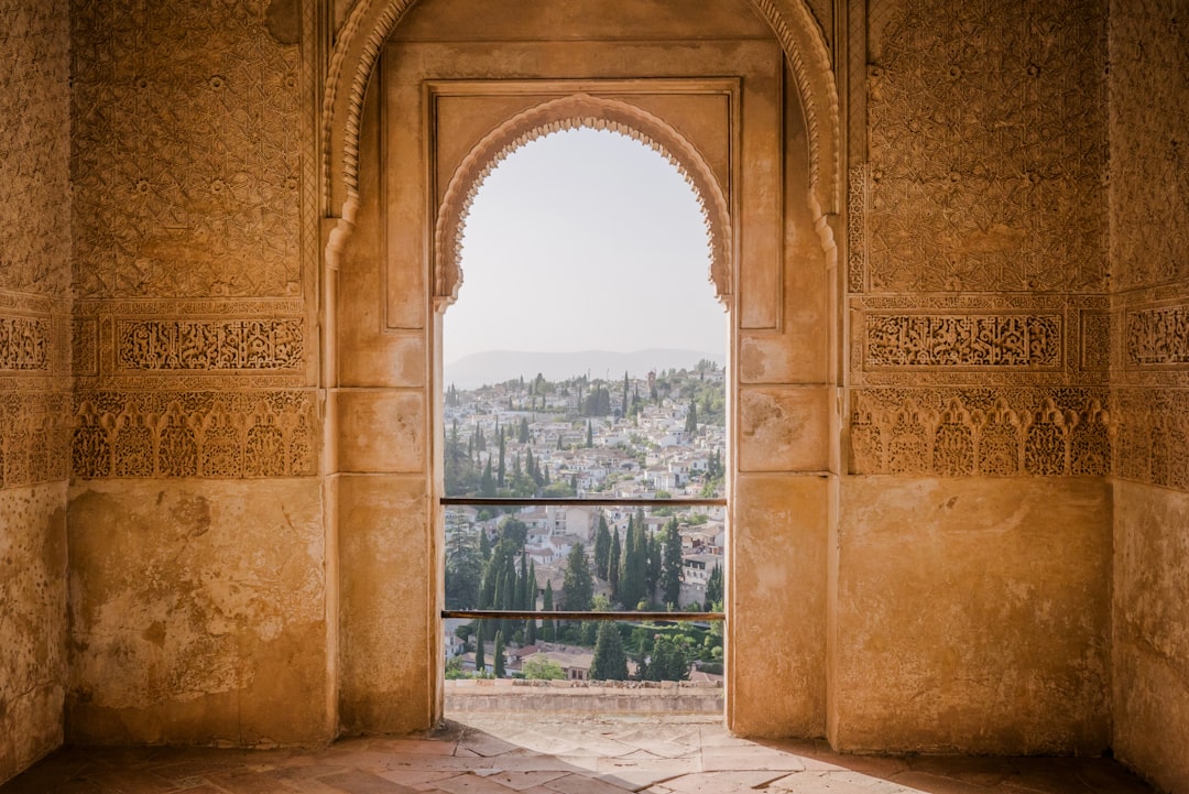 A view of the city through an arch in Alhambra, with intricate patterns on the wall and soft sunlight filtering from above. The window overlooks greenery and distant mountains under a clear sky. The patterns on the wall are in the style of the intricate designs found in Alhambra. –ar 128:85