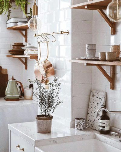 A photo of an elegant and modern kitchen with white tiles, wooden shelves and vintage decor. The countertop is made from marble or granite, featuring small floral arrangements in vases. A hanging coat rack hangs above the sink, adorned with copper pots and pans. In front of it stands a coffee machine and mugs on display. There's also a pot plant sitting next to a French press for coffee. This space exudes warmth through natural light streaming in.