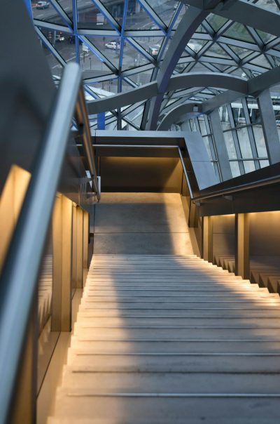 An architectural photograph of the interior staircase leading to an elevated platform in a busy public space. The steel structure has a glass roof above. The lighting is soft and natural, highlighting details like metalwork or glowing lights on walls. In front, there is another empty set of stairs leading down, adding depth to the scene. This perspective creates a sense of journey through different levels within the building, emphasizing its spaciousness and architecture in the style of an architectural photographer.