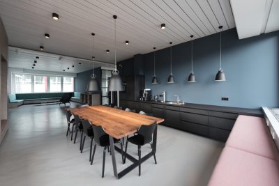 Modern kitchen with a long wooden dining table, black cabinets and a pink sofa in the background, a white floor, dark blue walls, ceiling lights hanging from pendant lamps, a grey industrial style interior design of a small apartment.