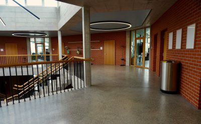 Ecofriendly interior design of the main hall in an educational building, featuring red brick walls and concrete floors with LED lighting to highlight energy efficiency. The space includes metal trash cans for sustainable waste management. A staircase leads up from the lower floor level towards upper balconies, offering panoramic views over students' activities during class hours. In one corner, a medium-sized poster display rack is set against a wall displaying information about environmental impact in the style of minimal editing.