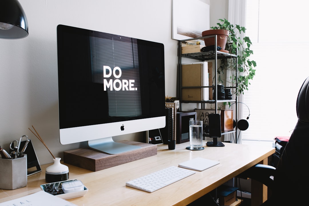 A minimalist desk with an iMac displaying the words “DORHemore.” The office is well-organized and has natural light, creating a calm work environment. A small plant sits on one side of the monitor, adding to its fresh atmosphere. –ar 128:85