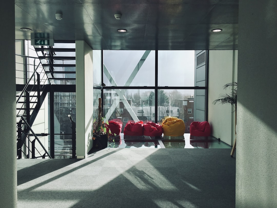 modern interior of an office building with large windows, bean bag chairs in red and yellow tones on the floor against one wall, gray carpet, staircase leading from above, sunlight falls through a big glass window onto the grey concrete walls, a shadow falls across part of the furniture and stairs, photo taken in the style of Canon EOS, 50mm lens –ar 4:3