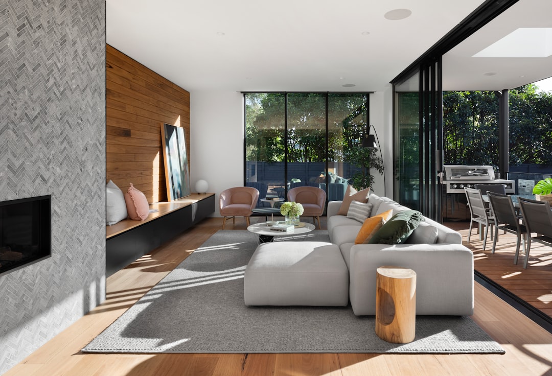 A modern home in Melbourne with an open plan living room, featuring white walls and light grey flooring. The focal point of the space is a large window that looks out onto trees, offering natural lighting into the interior. A fireplace sits on one side of the sofa area, adding warmth to the contemporary design elements such as the use of wood grain accents throughout the house. In front of it lies a wooden stool decorated with colorful cushions, creating a cozy atmosphere within the space.