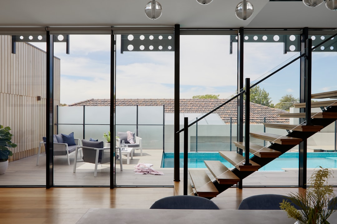 Wide shot of modern glass b-sized sliding doors leading to an outdoor pool area, in the background is an open living room with white walls and timber floorboards, grey colored accents. There is some mid-century furniture around the space. On one side you can see a balcony that has been replaced by black steel stairs leading down into a small swimming pool area. Shot taken from inside looking out through the windows, in the style of Hasselblad camera. –ar 128:85