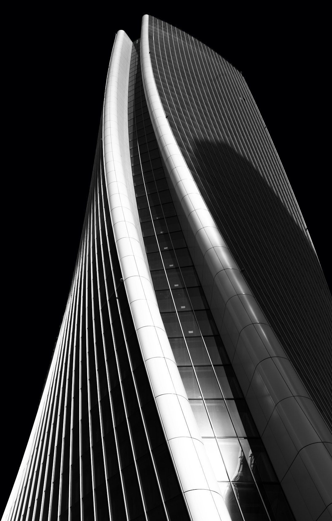 Black and white photography of the outside of a skyscraper in Milan, featuring modern architecture with modern curves and a steel structure. The building appears elegant and sleek with straight lines against a black background with dramatic lighting, showing architectural details in a high resolution, high contrast photograph. The photograph is award-winning and was taken in the style of HDR photography. –ar 81:128