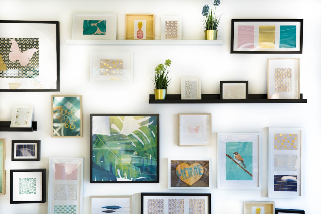 A white wall with various picture frames and art prints, showcasing different sizes of frame designs for display on the walls. The scene includes shelves holding framed artwork, with decorative elements like plants or potted flowers in small pots placed between some of them, adding color to the space. There is an emphasis on light colors such as pastel blues, greens, yellows, creating a fresh, modern look that adds charm and character to any room decor. Some of the artwork is displayed in the style of known artists. –ar 128:85