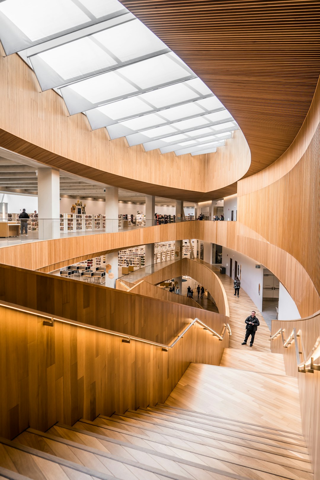 A photo of the interior design of a wood clad library and staircase in bright light from skylight windows in the style of [John Pawson](https://goo.gl/search?artist%20John%20Pawson). People are walking around curved wooden staircases with handles. White books are on shelves in the busy space with soft, warm lighting. It is architectural photography.