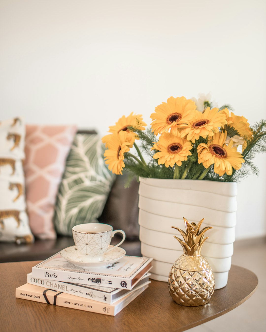 a white ceramic vase with yellow gerberas, next to it is an elegant gold pineapple and two books on the coffee table, in front of sofa with pink boho pillow, interior design aesthetic photography style