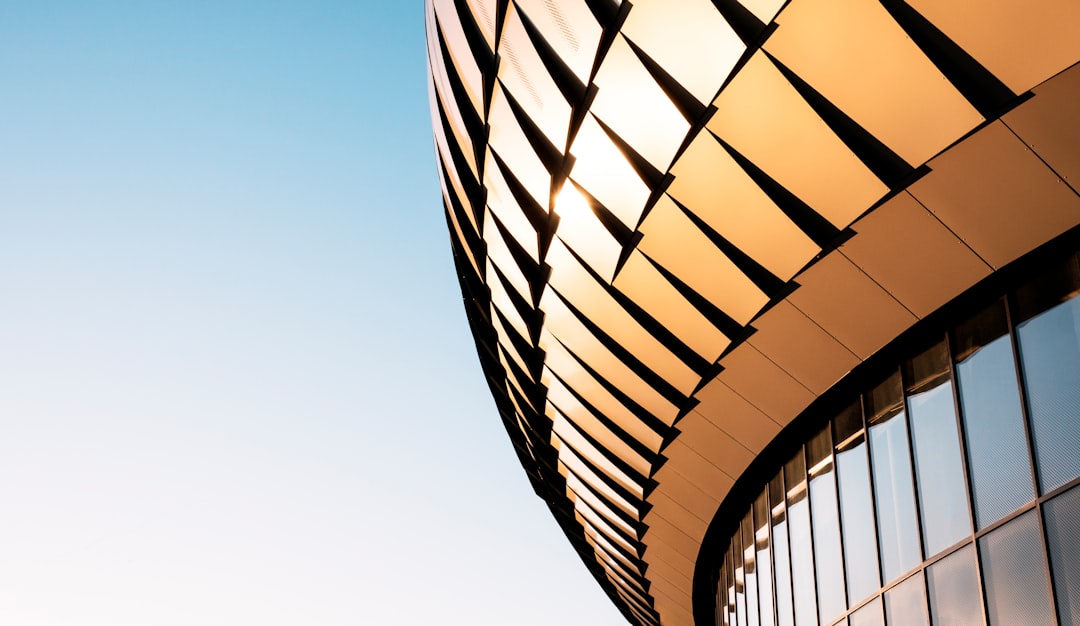 Close up photo of a curved modern building with metal cladding, featuring a light gold and black color palette against a blue sky background with cinematic lighting, captured in the style of Canon EOS R5. –ar 64:37