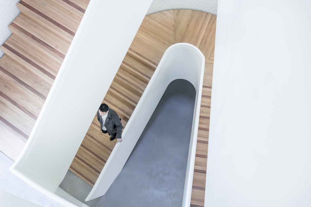 Aerial shot of an Asian man in a suit walking on the curved staircase, with wood grain and a gray floor, white walls, in the style of modern architecture, interior design photography, natural light, minimalism, in the style of Jimmy Nelson