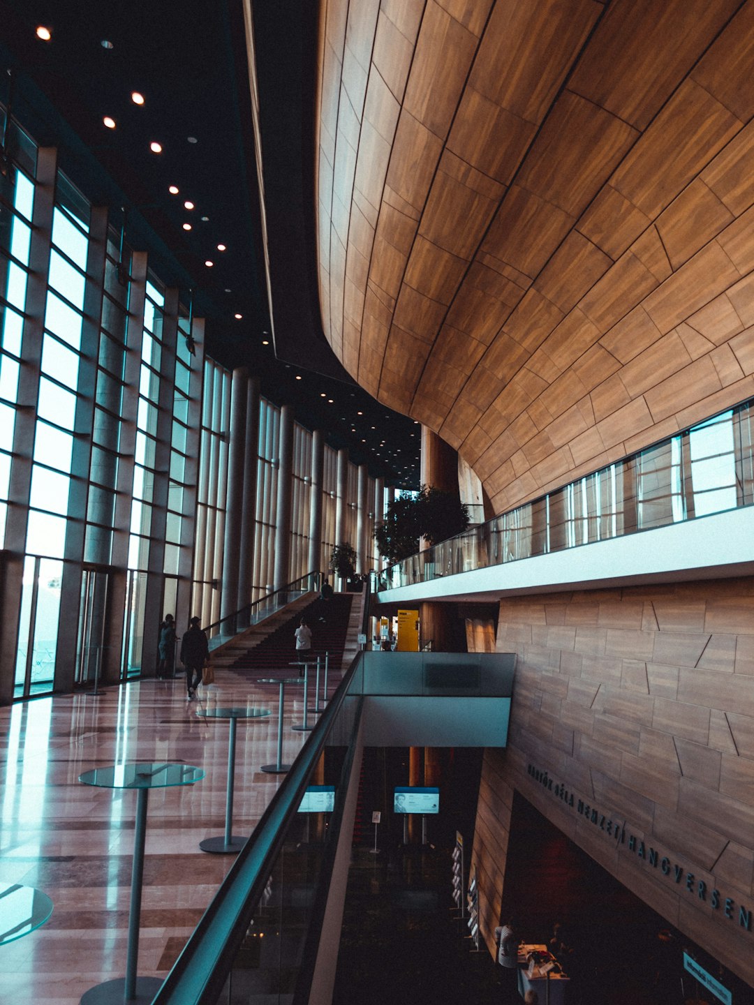 The interior of the concert hall features a wooden ceiling and walls, with large glass windows on one side that provide stunning views outside. The main entrance is located in front, featuring an elevated staircase leading to its upper level seating area. People can be seen walking around inside or sitting at various chairs enjoying music or showcasing their dance skills. In a sharp focus photography style. –ar 3:4