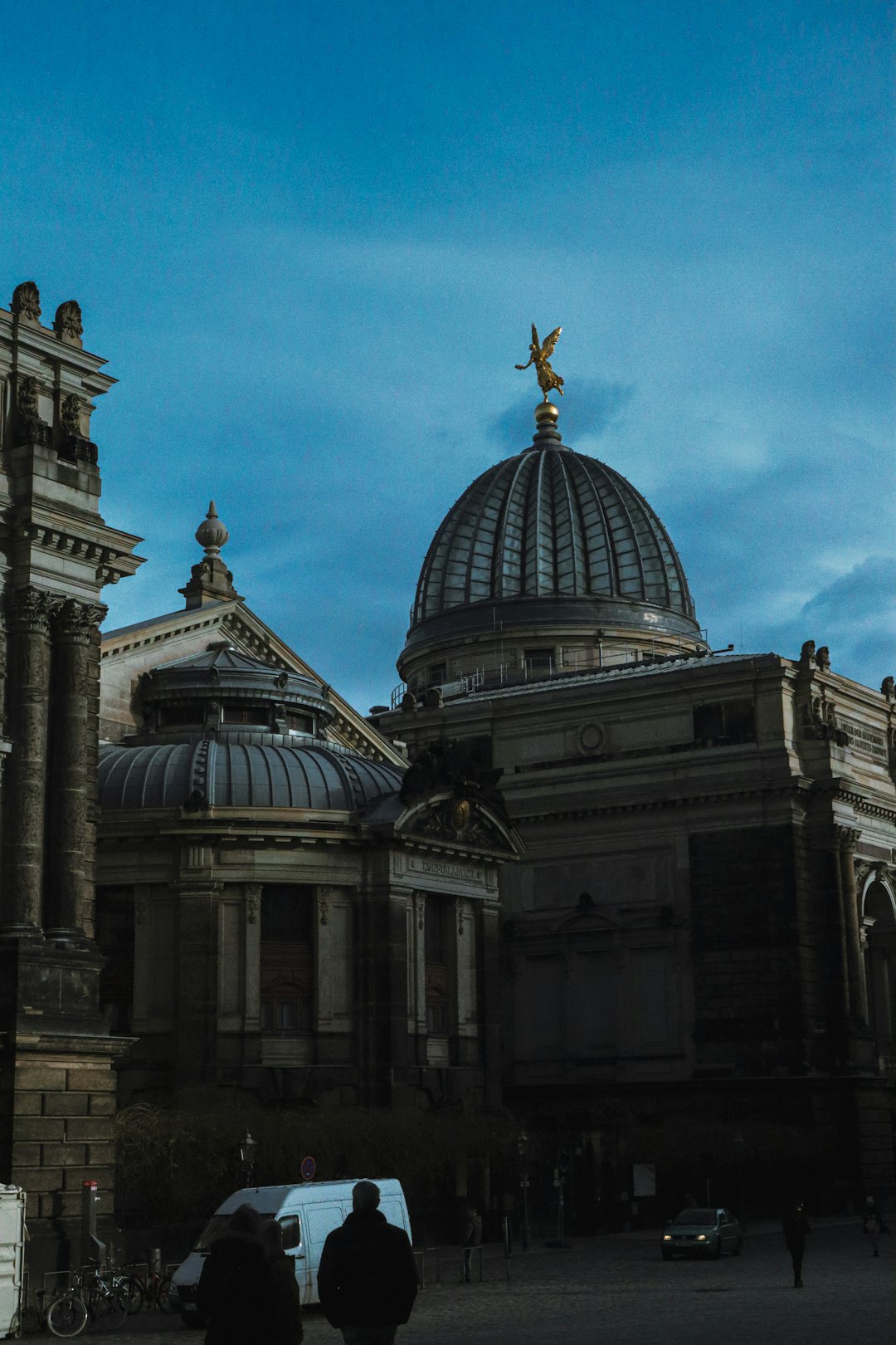 photo of the back side of an old art museum in downtown, at blue hour with an angel statue on top of its dome, with people walking around and some white van parked nearby, captured using a Fujifilm XT4 and Sony FE2350mm f/2.8 G OSS lens in the style of G OSS. –ar 85:128