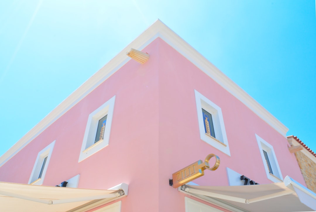 pink building in the style of Greek architecture, blue sky, sun shining on it, white roof and window frames, with golden decoration. High quality editorial photography with professional color grading of a natural light magazine shoot on a bright sunny day. –ar 64:43