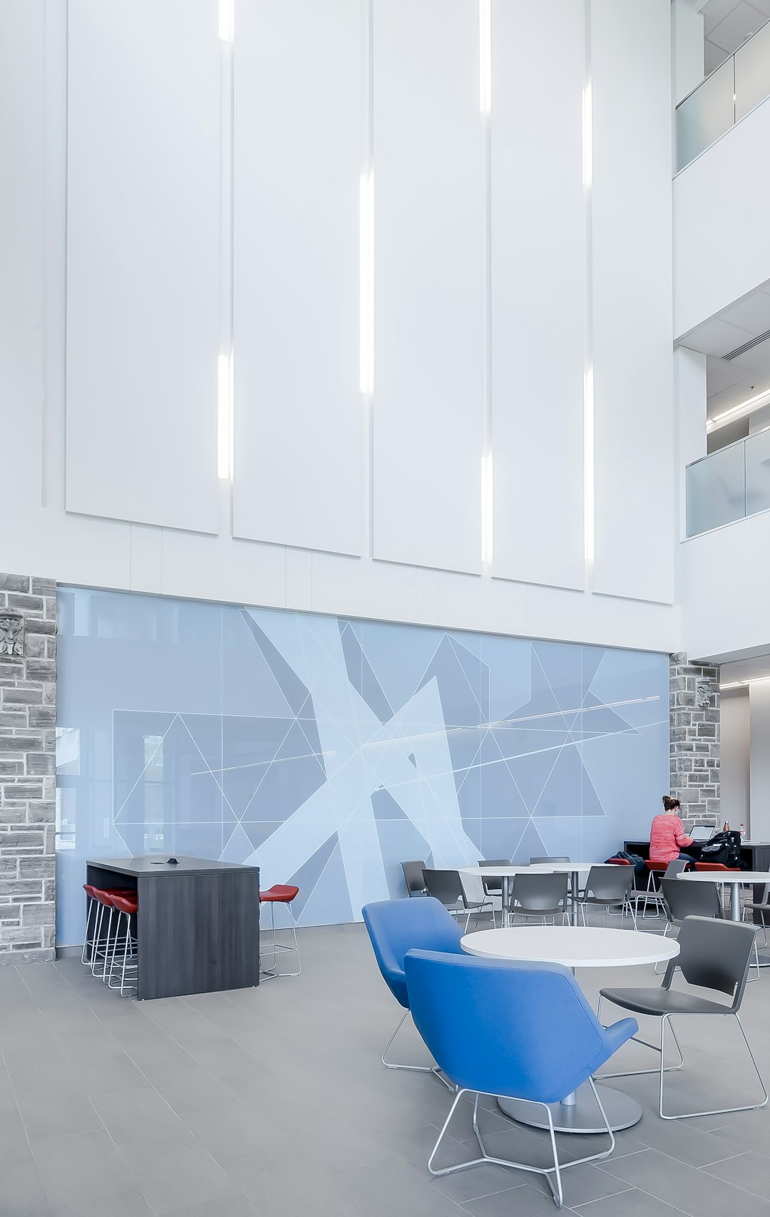 A photo of an interior wall in the lobby at Queen’s University, featuring three large white accent panels with blue geometric patterns. The bright and airy space showcases modern furniture like chairs around tables for students to socialize or study. In one corner there’s even a beehive shaped bar table for drinking coffee while chatting. A small group sits scattered across some seating area nearby.