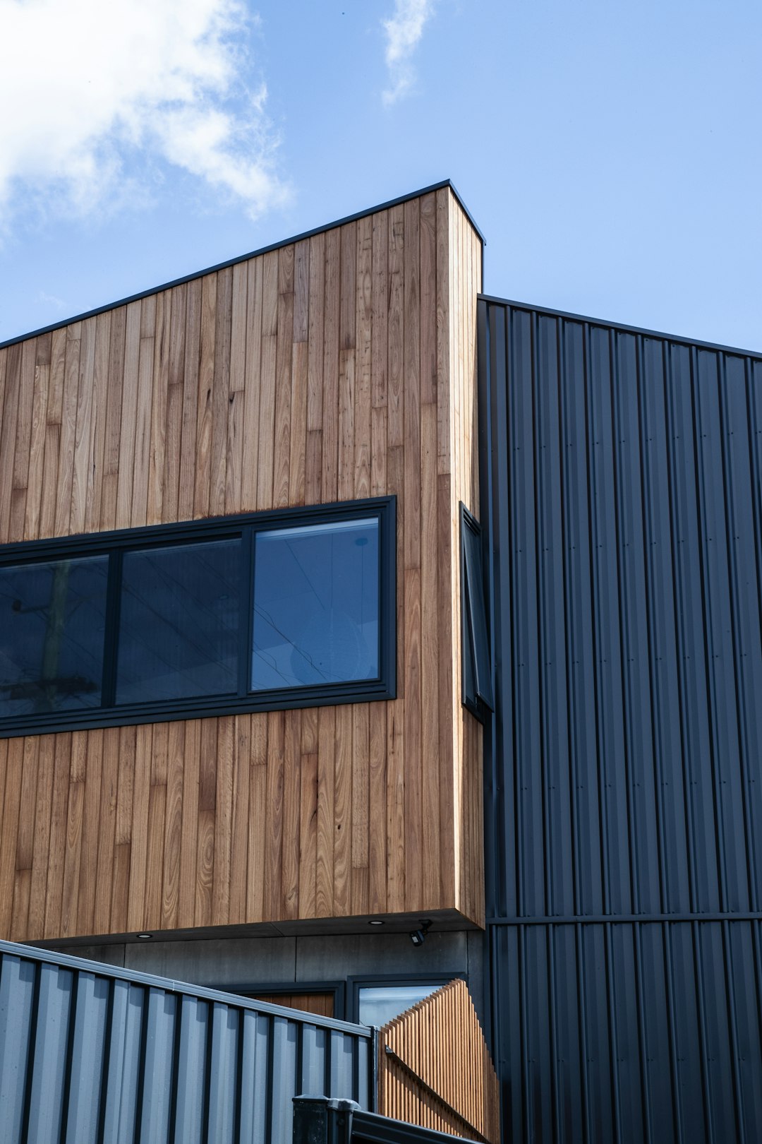 A close up photo of the corner exterior wall of an industrial building made from timber cladding and black iron accents, under blue sky, wooden slats on one side, flat roof with black eaves, black window frames. The house is in New Zealand, the building has two stories. Photo taken at eye level. –ar 85:128