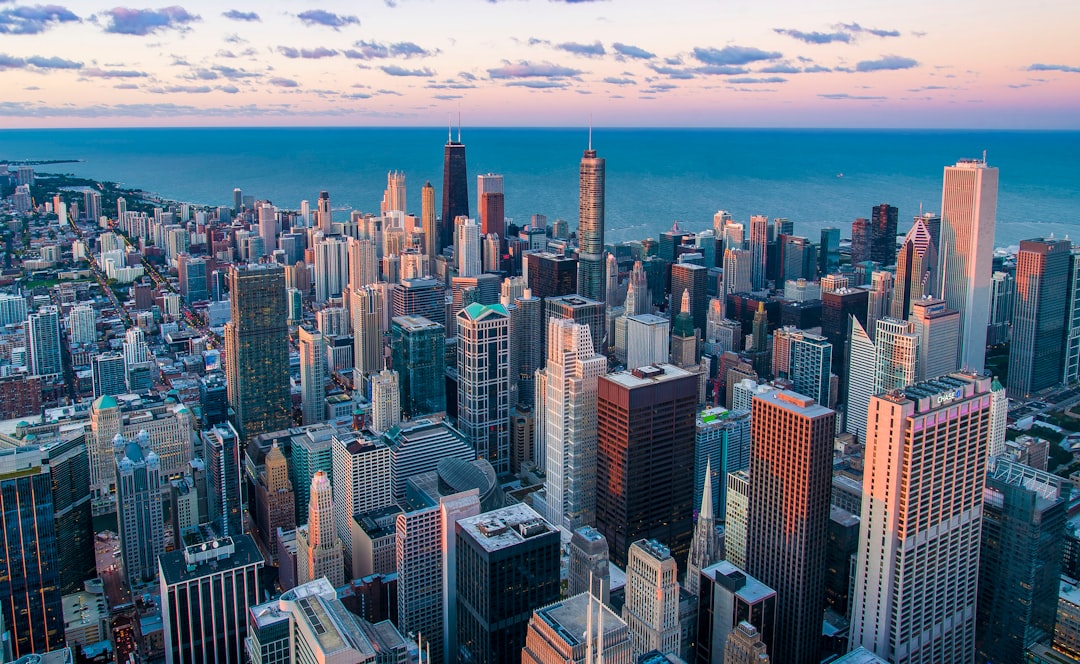 Aerial view of the Chicago skyline at sunset, with skyscrapers and buildings in various shades of gray, white, blue, pink, purple, and orange against an ocean backdrop. The cityscape is bustling during golden hour, with warm sunlight casting long shadows over the urban landscape. Shot on a Canon EOS R5 camera with an RF 24-70mm f/8 lens.