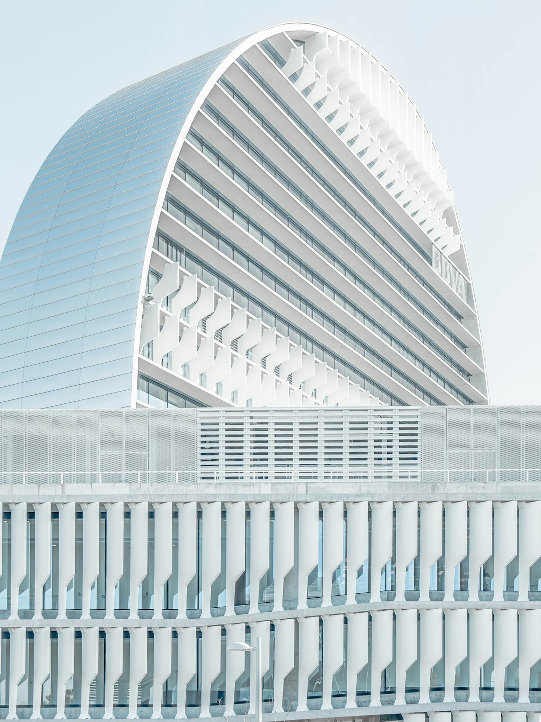 A modernist building with white walls and large windows, the roof is curved like an arch made of horizontal slats that create shadows on its surface, with a minimalist architectural style. The background color has light blue tones, creating a sense of calmness and simplicity. In front of it stands another highrise office building with a glass curtain wall.