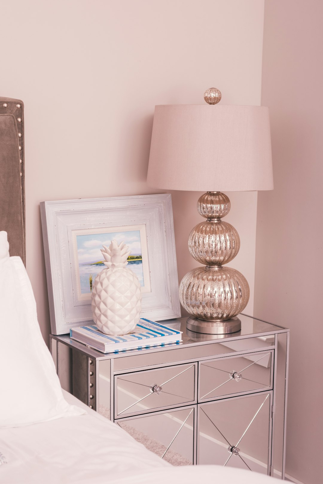 a close up photo of an elegant nightstand with silver mirror finish, a large white framed picture on the top and a glass lamp in light pink next to it, a small pine apple is placed beside them. The background color should be pastel pink walls. A stack of magazines sits under one corner of that night stand. This scene embodies modern luxury interior design style. –ar 85:128