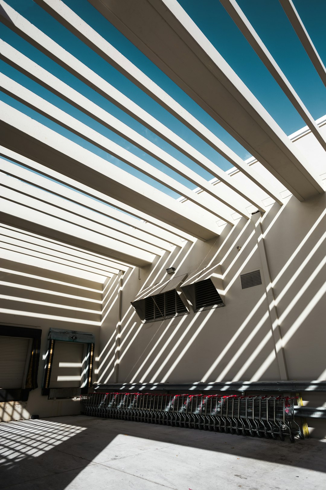 A photograph of the exterior ceiling and roof of an outdoor shopping center, featuring white lines that create geometric patterns on its surface. The sunlight casts dramatic shadows through these stripes onto two rows of shopping carts placed below. This shot captures the modern architecture with its clean design, emphasizing light effects and creating visual interest in architectural details. The photograph was taken with a Fujifilm GFX 50S camera using settings for high detail and with a wide angle lens perspective.