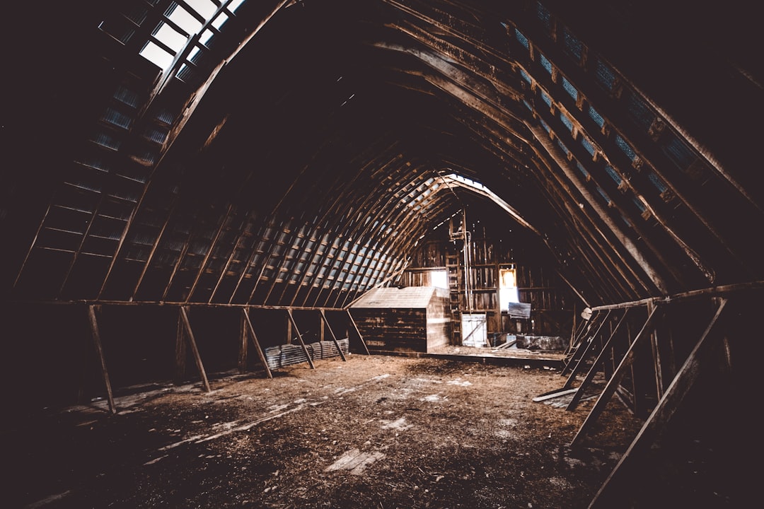 A dark, abandoned barn interior with wooden beams and a central opening leading to the attic area. The ceiling is slanted in an arch shape. There are construction materials scattered around, indicating work being done or repair works taking place. A faint light from outside shines through one of the skylights on top. Shot in the style of Canon EOS R5 camera using a wideangle lens. –ar 128:85