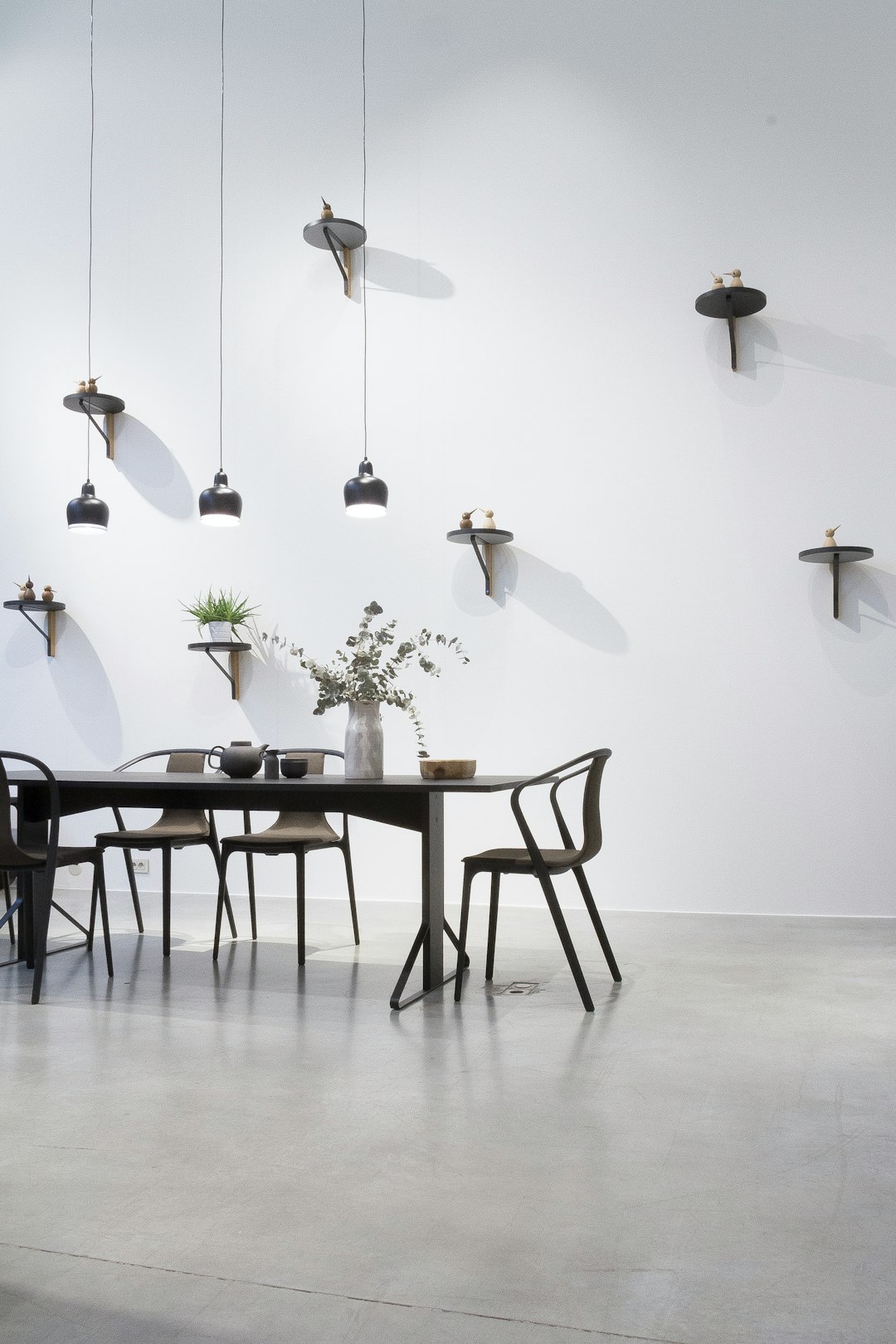 A dining table and chairs are set against white walls, with black metal wall lamps hanging on the left side. The dark wood tabletop sits above an indoor concrete floor. Small vases or flower arrangements sit on one end of each chair. A black wooden shelf hangs above in front of two rectangular lights that cast long shadows across the room. No other furniture is around the tables. –ar 85:128