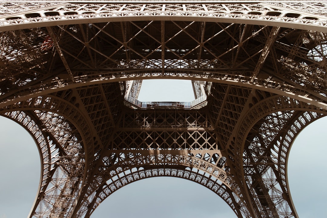 A closeup shot of the intricate structure and details beneath one side, showcasing how it’s made up of interlocking steel beams and arches that form an oval shape at its base, with visible gears moving in motion. The background is clear sky against the Eiffel Tower. –ar 128:85