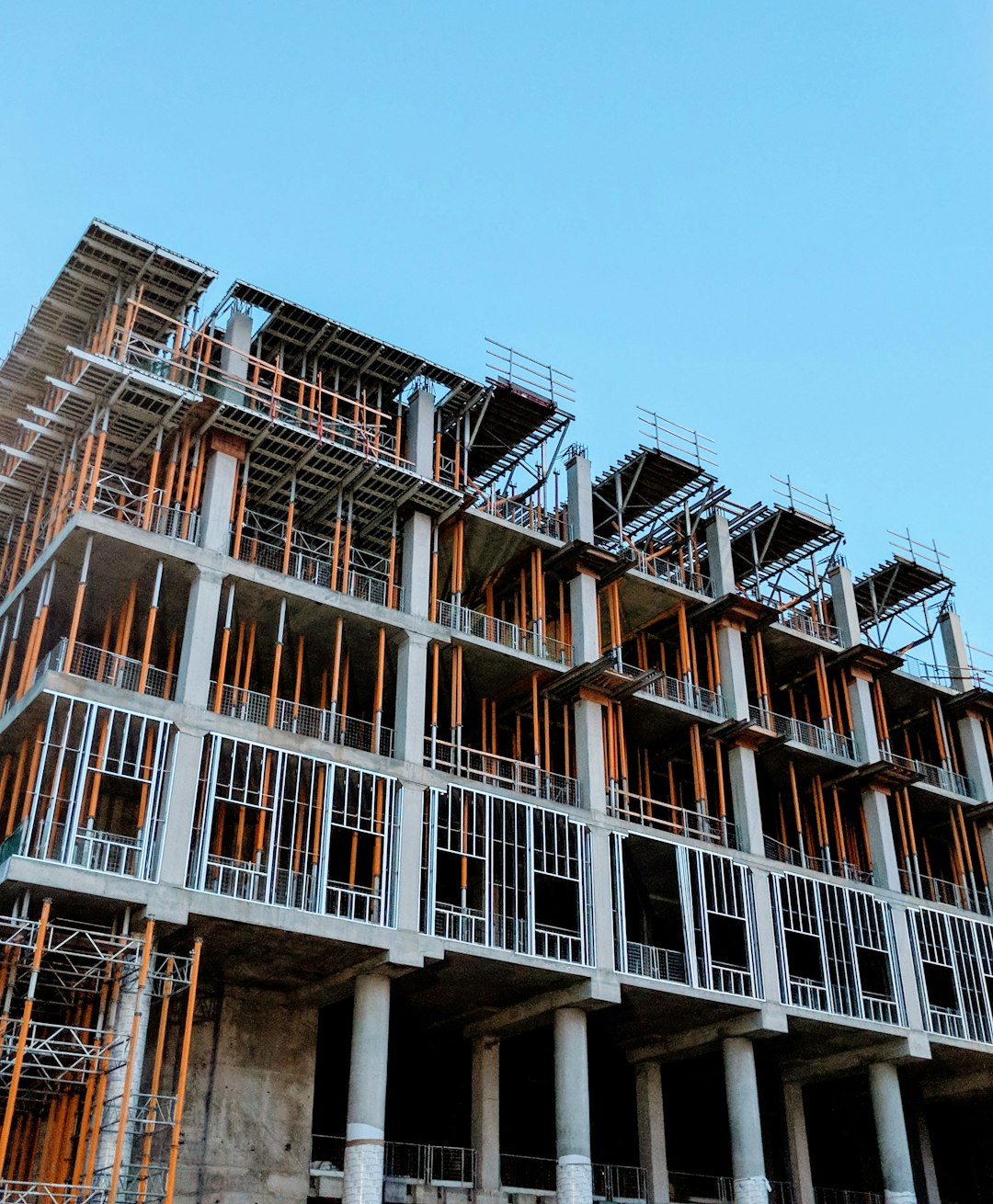 A modern building is being built with many columns and steel beams, while the construction site has orange scaffolding and blue sky in the background. The structure of the tall concrete multistory apartment complex includes an elegant balcony on each floor. It’s covered in sunlight and there are no people around. The photography has an ultra realistic photo quality in the style of no specific artist. –ar 105:128