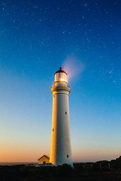 A white lighthouse shines brightly against the night sky, illuminating its surroundings with warm light. The stars twinkle in the clear blue evening sky above it. The photograph is in the style of realistic photography.