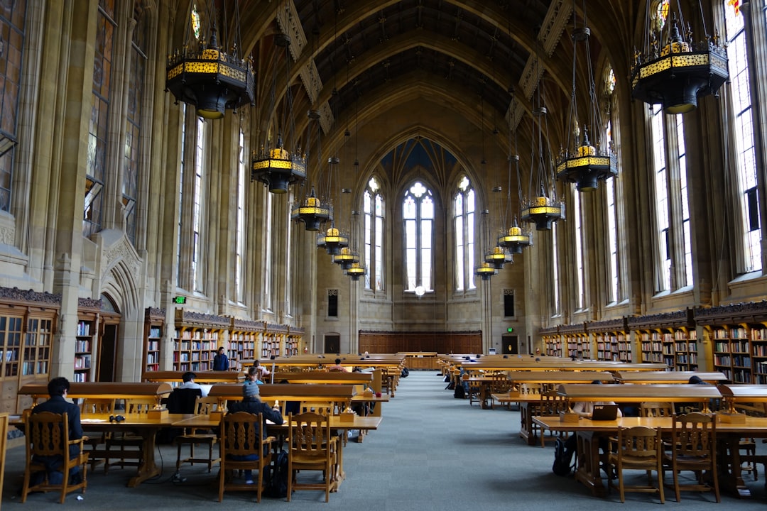 The fields library at the University of Washington has many tables and chairs with students studying in them. The ornate gothic architecture features beautiful large windows and tall ceilings. Many hanging lights illuminate the library.