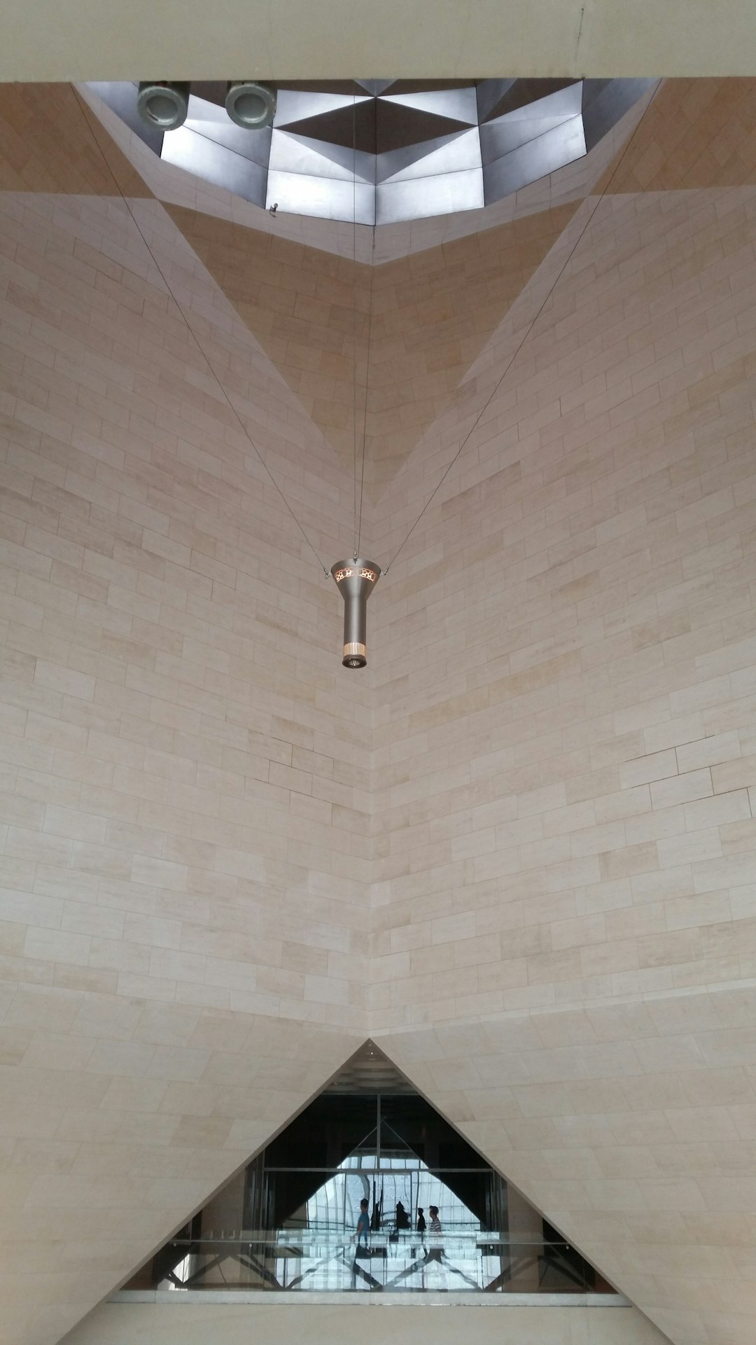 The National Museum of Palestine in Doha has beige stone walls. The ceiling is suspended with thin metal cables. A large skylight is above the entrance. On the ground floor, a glass window looks into the space. –ar 9:16