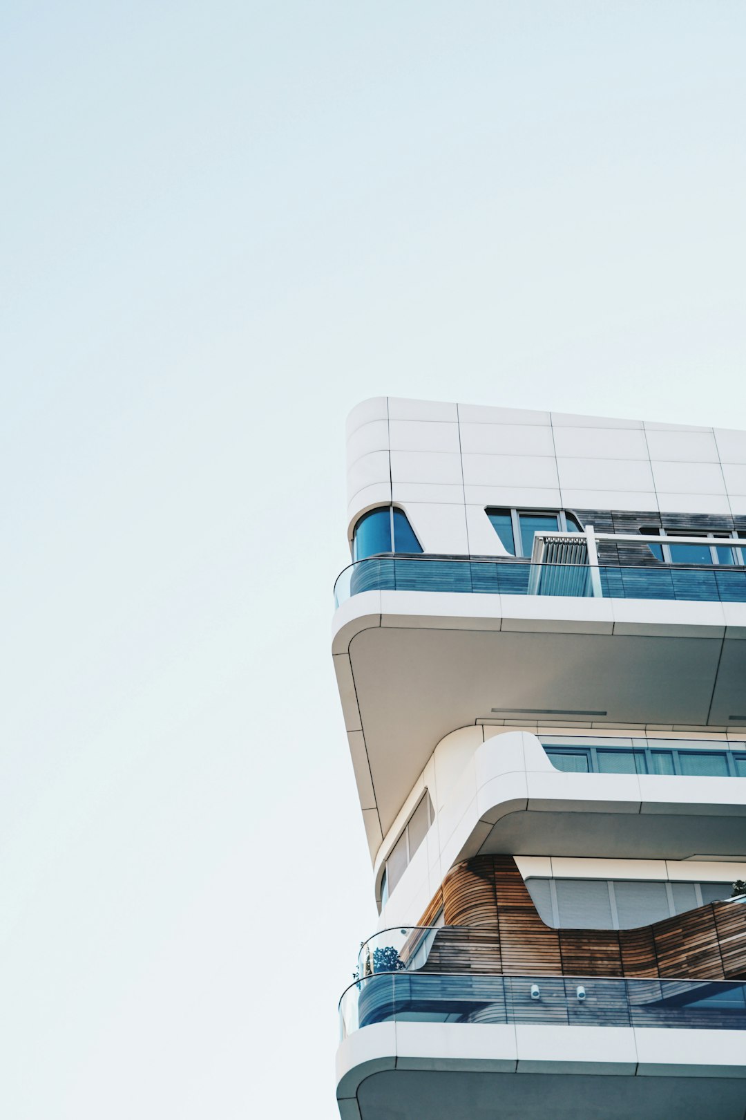 A modern building with curved balconies, white and blue color scheme, against the clear sky. The balcony of each floor is decorated in wood, creating an elegant urban landscape. High resolution photography with color grading, soft shadows and clean sharp focus in natural light, presented in the style of high quality digital photography. Shot in the style of Hasselblad X2D. –ar 85:128