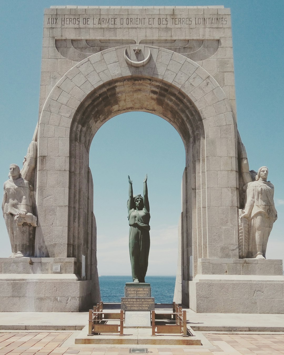 A monumental stone archway, flanked by statues of a woman and man holding the French flag in one hand above their head with “Hommage aux héros de la marine et des États-Unis” written on it, stands against an ocean backdrop. The scene is captured from behind the monument, with the sea visible through its arched opening. A serene blue sky serves as the background, enhancing the grandeur of the structure’s architecture. –ar 51:64