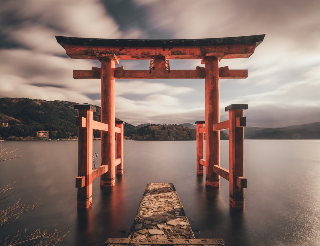 A red torii gate standing on the edge of an ancient Japanese lake, surrounded by mountains and clouds. The scene is captured in long exposure photography style with soft lighting and a tranquil atmosphere.