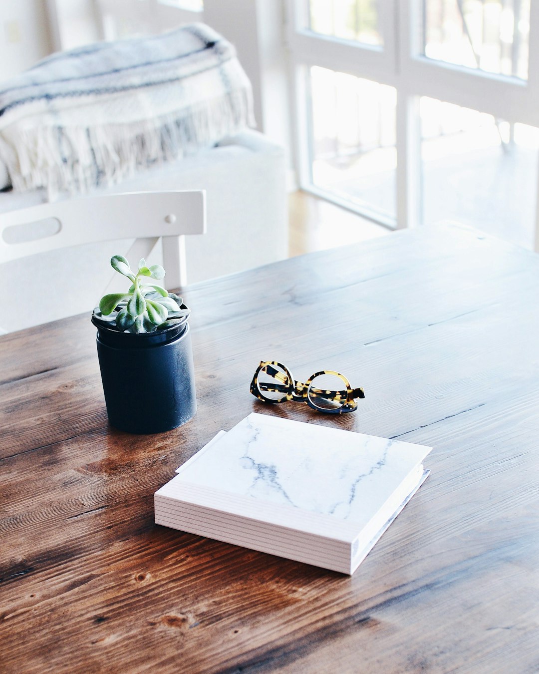 A photo of white marble A5 notebook on the coffee table, next to it is sunglasses and small black plant pot with succulent in front of window with natural light, boho style living room interior design –ar 51:64